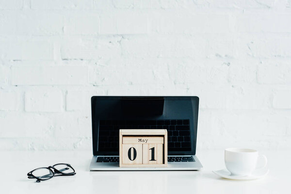 Wooden calendar on laptop with blank screen, eyeglasses and cup 