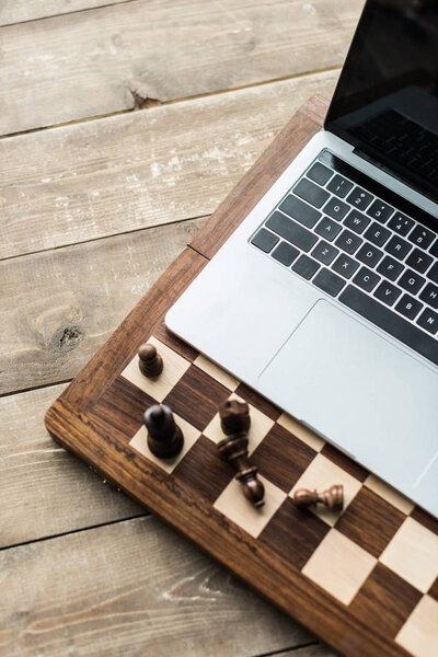 Cropped image of chess board with chess pieces and laptop on rustic wooden surface