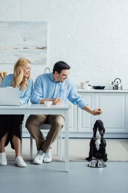 Man and woman drinking coffee and playing with their dog by kitchen table with laptop clipart