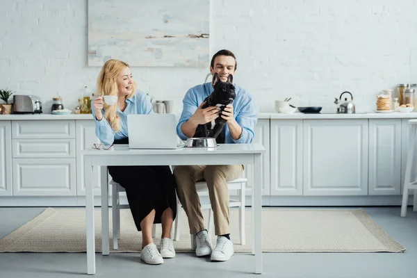 男人和女人喝咖啡 玩法国斗牛犬在厨房桌子上的笔记本电脑 — 图库照片