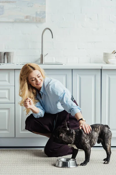 Χαμογελώντας Χαϊδεύοντας Frenchie Σκυλί Ξανθιά Γυναίκα Στο Πάτωμα Της Κουζίνας — Φωτογραφία Αρχείου