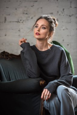 attractive brunette woman in trendy jumper sitting on armchair and looking away
