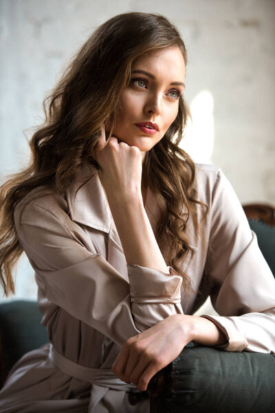 brunette woman with long hair in beige trench coat posing in armchair