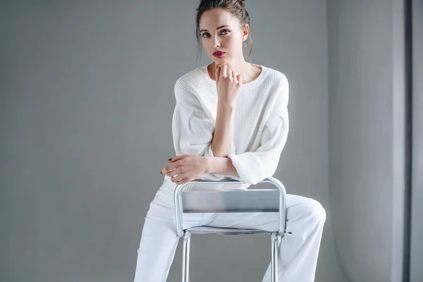 流行の白服の椅子に座っているとグレーのカメラ目線で美しいブルネットの女性の肖像画 — ストック写真