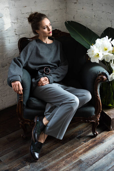 full length view of elegant young woman sitting on armchair and looking away