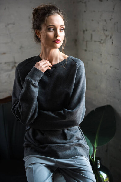 portrait of beautiful brunette woman in trendy grey clothes sitting on armchair and looking away