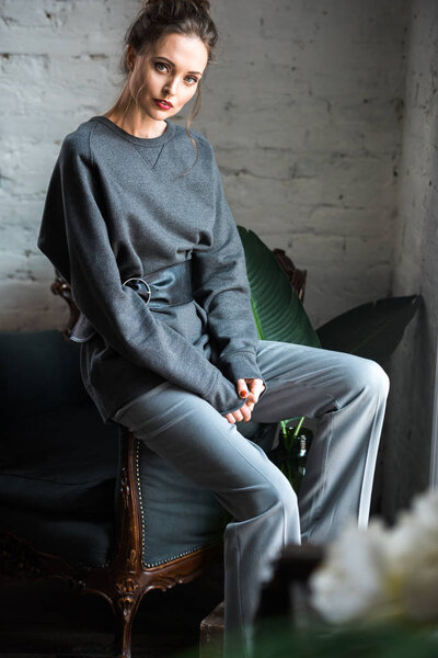 beautiful young woman in trendy grey clothes sitting on armchair and looking at camera