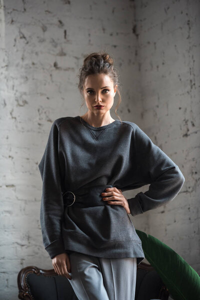 portrait of beautiful young woman in trendy jumper with belt standing with hand on waist and looking at camera