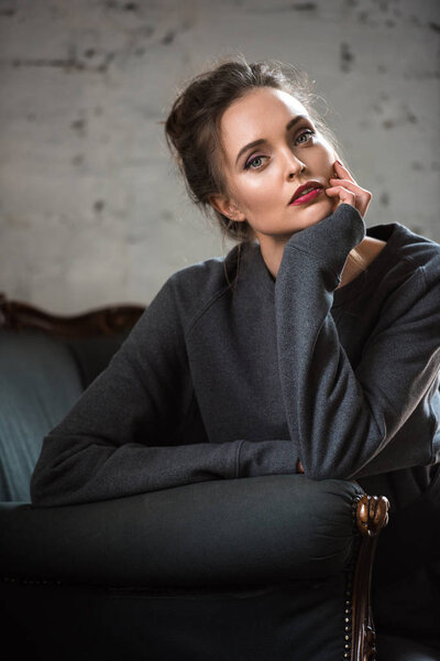 attractive brunette woman in trendy jumper sitting on armchair and looking at camera 