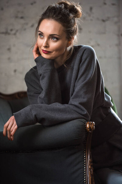 portrait of pensive brunette woman in grey clothes sitting on armchair and looking away