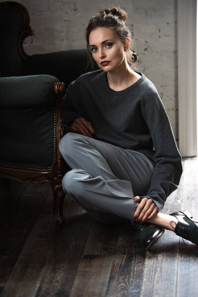 beautiful brunette woman in stylish grey clothes sitting on hardwood floor and looking at camera