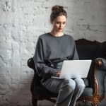 Schöne brünette Frau in stilvollem Outfit mit Laptop, während sie auf einem Sessel sitzt