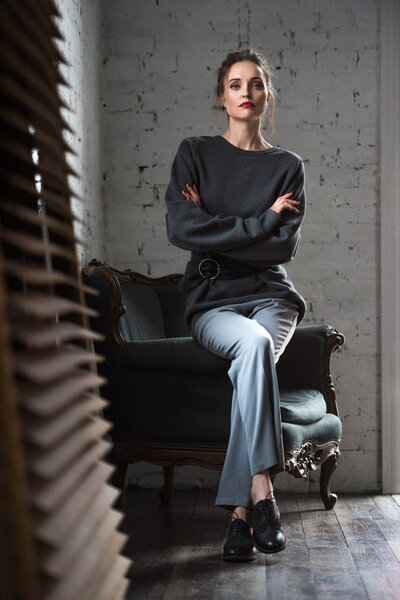 full length view of stylish young woman with crossed arms sitting on armchair and looking at camera