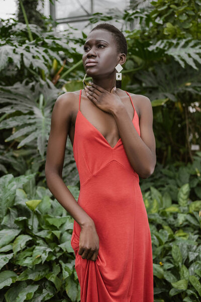 fashionable african american girl with short hair posing in trendy red dress in tropical garden