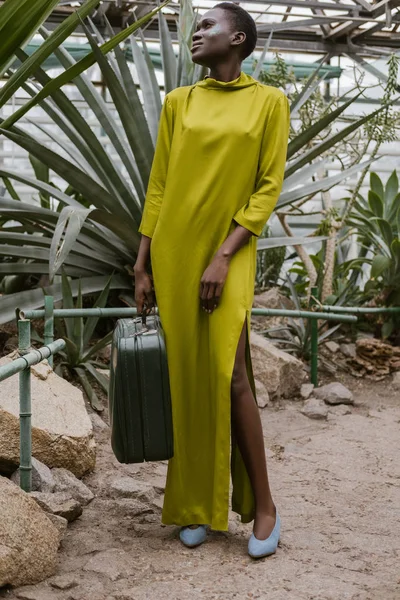 Стильная Африканская Американка Желтом Платье Позирует Дорожной Сумкой Тропической Оранжерее — стоковое фото