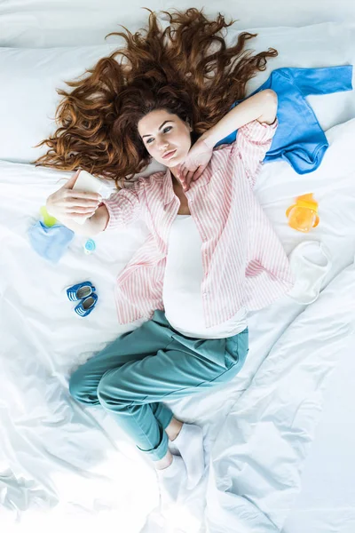 ベビー服の中でベッドで Selfie を取って妊娠中の女性のトップ ビュー  — 無料ストックフォト