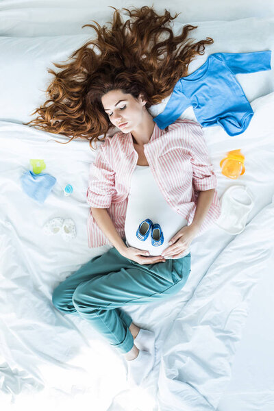 Top view of pregnant woman in bed sleeping among baby clothes