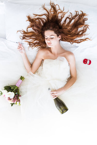 Top view of woman wearing wedding dress lying in bed with champagne and wedding rings