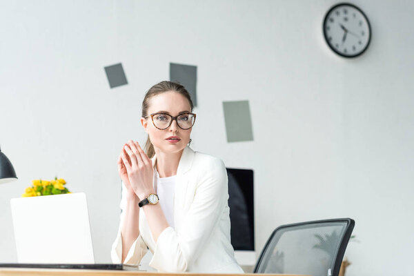 портрет деловой женщины в белом костюме и очках на рабочем месте с ноутбуком в офисе
