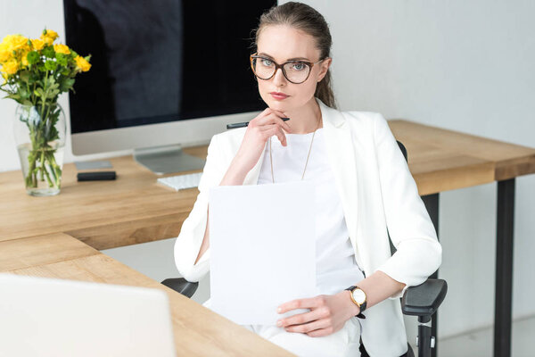 portrait of businesswoman in eyeglasses with documents at workplace with laptop in office