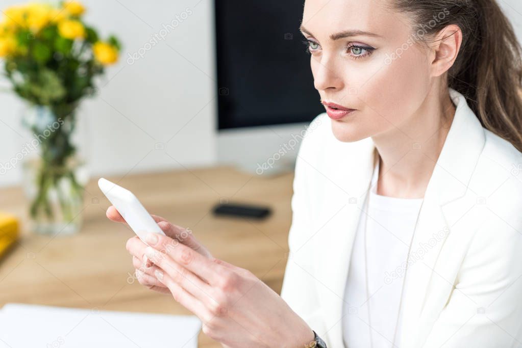 selective focus of pensive businesswoman in stylish suit with smartphone in office