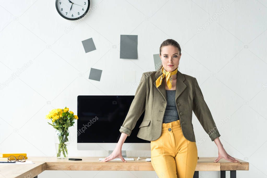beautiful businesswoman in fashionable clothing leaning on workplace with computer screen and bouquet of flowers in vase in office