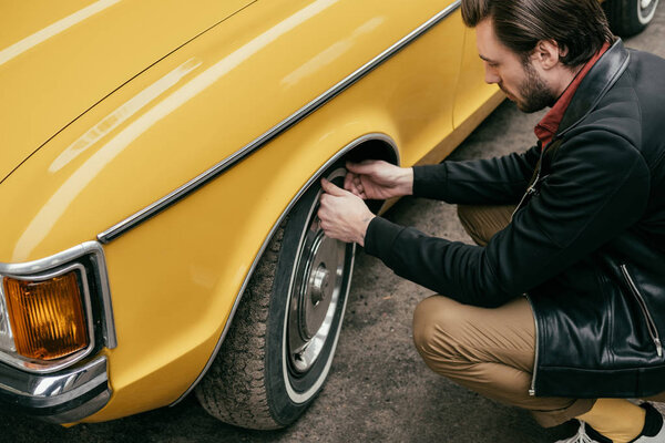 cropped shot of handsome man in leather jacket fixing wheel of yellow retro car 