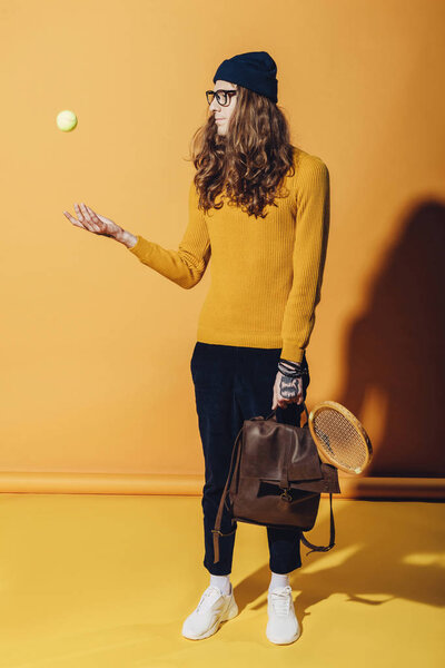 fashionable man with leather backpack and wooden racket throwing up tennis ball, on yellow