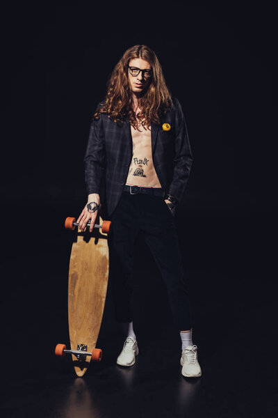 stylish tattooed skater posing with longboard, on black