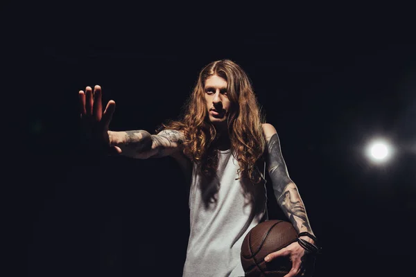 tattooed man with long hair gesturing and holding basketball ball, isolated on black with backlit