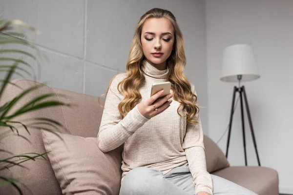 Schöne junge Frau mit Smartphone, während sie auf der gemütlichen Couch zu Hause witzelt — Stockfoto