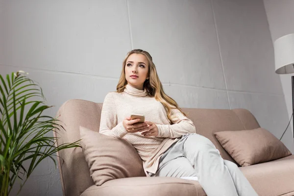 Jeune femme réfléchie utilisant un smartphone sur le canapé à la maison et détournant les yeux — Photo de stock
