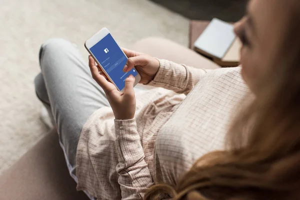 Обрезанный снимок женщины на диване с помощью смартфона с приложением facebook на экране — стоковое фото