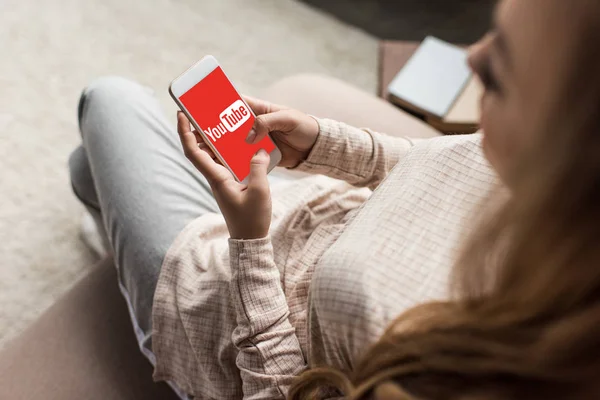 Cropped shot of woman on couch using smartphone with youtube logo on screen — Stock Photo