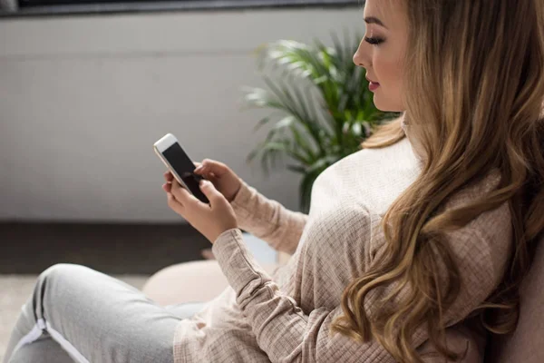 Attractive young woman using smartphone while sitting on couch at home — Stock Photo