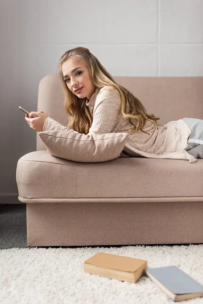 Attraente giovane donna utilizzando smartphone sul divano a casa e guardando la fotocamera — Foto stock