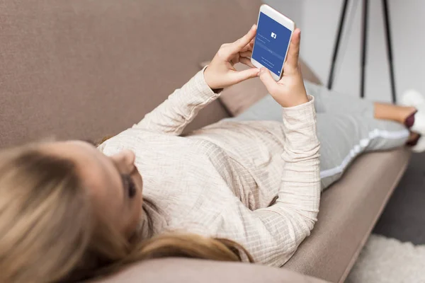 Femme sur le canapé en utilisant smartphone avec application facebook à l'écran — Photo de stock