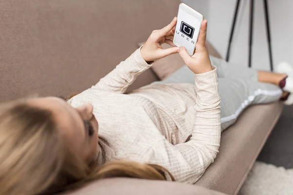Woman on couch using smartphone with uber logo on screen — Stock Photo