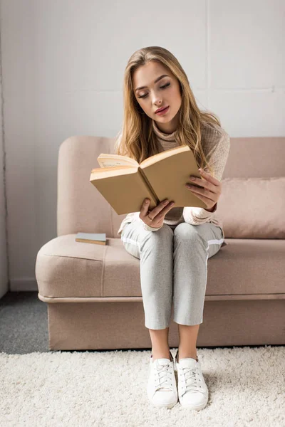 Mujer joven concentrada leyendo libro en el sofá acogedor en casa - foto de stock