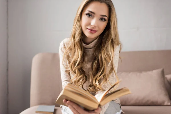 Belle jeune femme lecture livre sur canapé confortable à la maison — Photo de stock