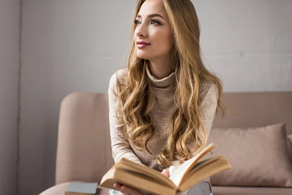 Thoughtful young woman with book on cozy couch at home — Stock Photo