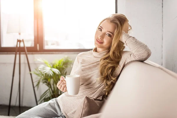 Belle jeune femme buvant du café sur le canapé à la maison — Photo de stock