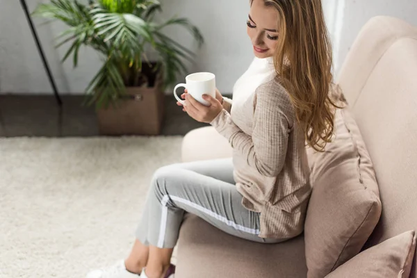 Bela jovem com xícara de bebida quente sentado no sofá em casa — Fotografia de Stock