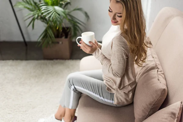 Beautiful young woman with cup of hot drink sitting on couch at home — Stock Photo