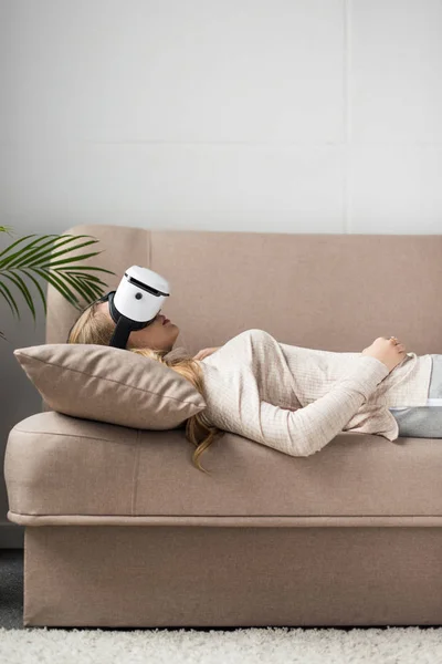 Vista lateral de la mujer joven en auriculares de realidad virtual tumbado en el sofá - foto de stock