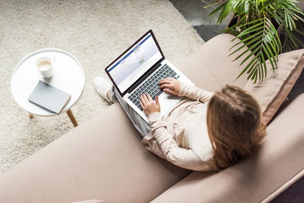 Vista de ángulo alto de la mujer en casa sentado en el sofá y el uso de la computadora portátil con el sitio web de facebook en la pantalla - foto de stock