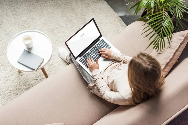Hochwinkel-Ansicht einer Frau zu Hause, die auf der Couch sitzt und Laptop mit Google-Suche auf dem Bildschirm benutzt — Stockfoto