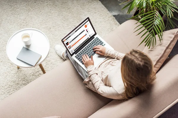 Vista de ángulo alto de la mujer en casa sentado en el sofá y el uso de la computadora portátil con el sitio web soundcloud en la pantalla - foto de stock