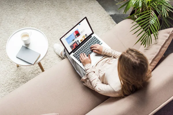 Vista de ángulo alto de la mujer en casa sentado en el sofá y el uso de la computadora portátil con el sitio web de ebay en la pantalla - foto de stock