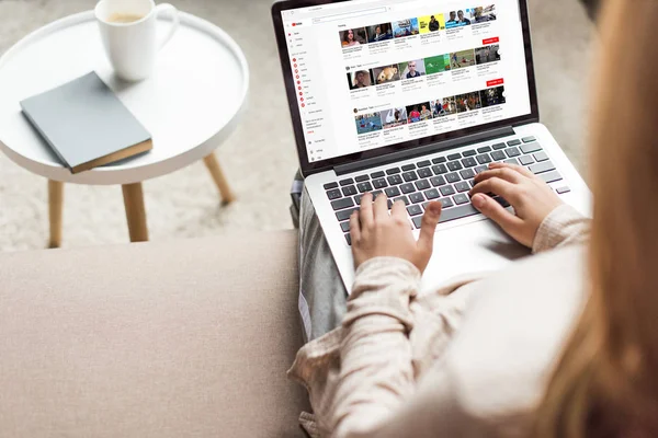 Corte tiro de mulher em casa sentado no sofá e usando laptop com site do youtube na tela — Fotografia de Stock